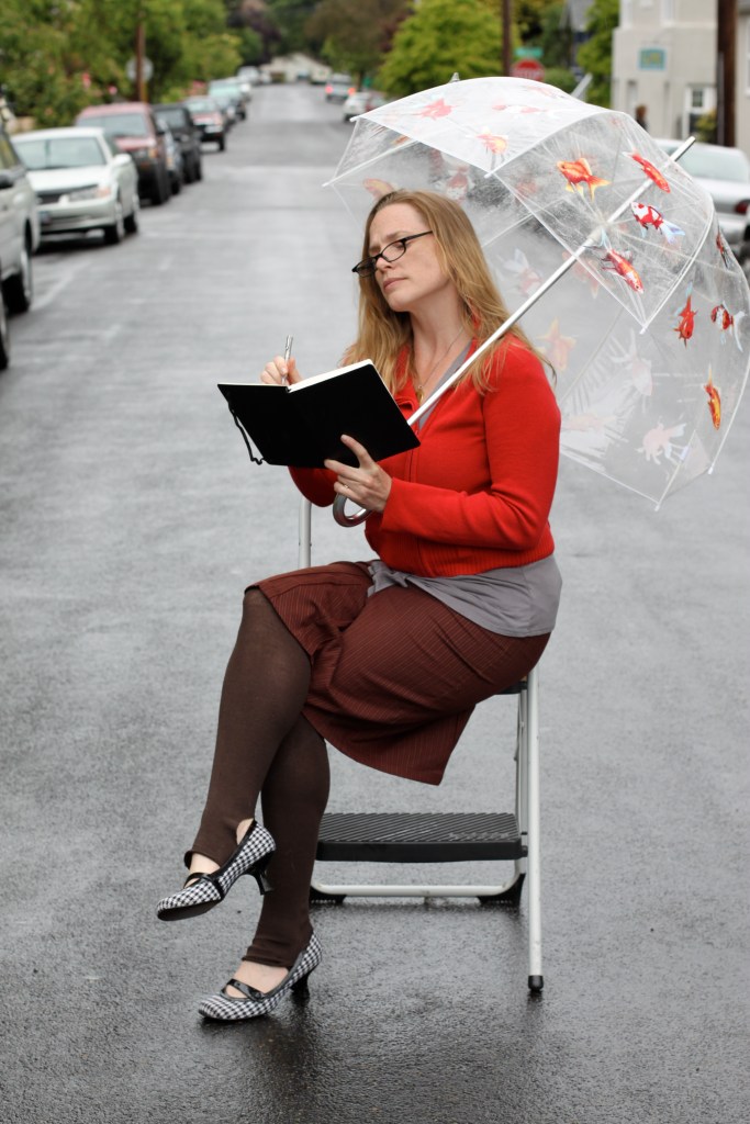 A woman in her early 30's with blonde  shoulder length hair is sitting on a chair in the middle of a road. She  wears glasses and is dressed in a bright red sweater, a brown knee length skirt, and brown leggings. Her small heel shoes are black and white houndstooth. She is writing in a black notebook as she holds an umbrella over her shoulder.