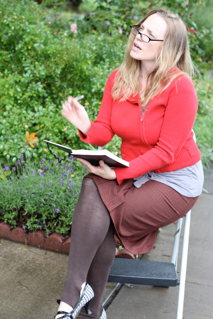 A woman in her early 30's with blonde  shoulder length hair is sitting on a chair in next to a garden. She wears glasses and is dressed in a bright red sweater, a brown knee length skirt, and brown leggings. Her small heel shoes are black and white houndstooth. She looks away from the camera speaking to someone out of the frame. She holds a notebook and a pen.