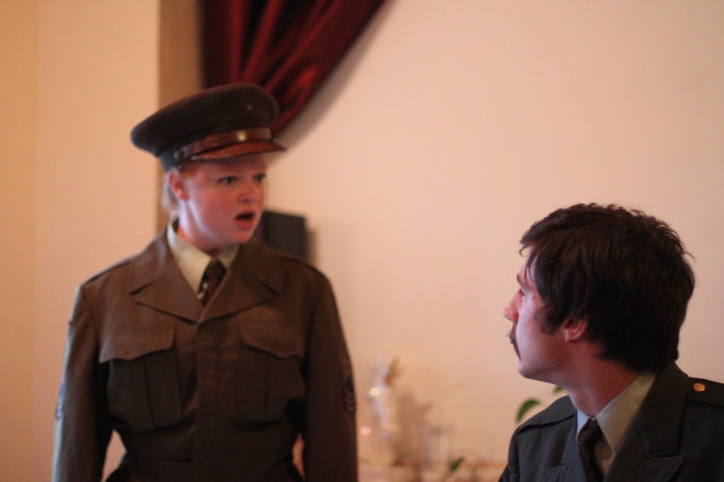 A young woman in a 1940's military uniform looks shocked as she speaks with a male officer whose face is turned away from the camera.