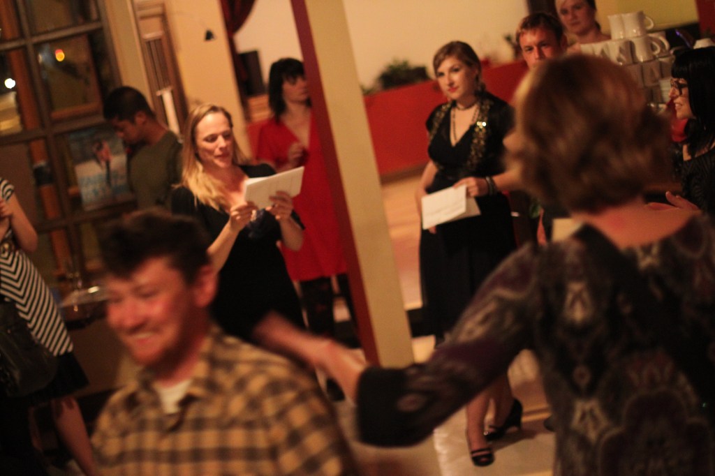A group of people stand around a woman who is reading a paper.