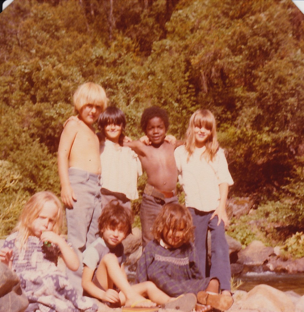 A group of seven kids posing in front of a river. Four kids are standing and they all have their arms around each other. From left to right: a blonde, white, boy, topless and wearing blue jeans; a dark haired, Latina girl with a white blouse; a black boy with short hair is topless in blue jeans, and a white girl with shoulder length blonde hair in a white blouse and blue jeans. Three younger children are sitting on the rocks. From left to right: a white blode girl in a dress blowing a balloon, a dark haired boy with an olive complexion in a blue shirt and shorts, and a Mexican girl with curly hair covering her face, she is dressed in a dress. 