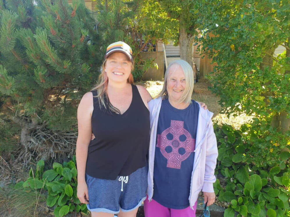 A middle aged white woman and a white elderly woman pose smiling in front of green bushes. They are family.