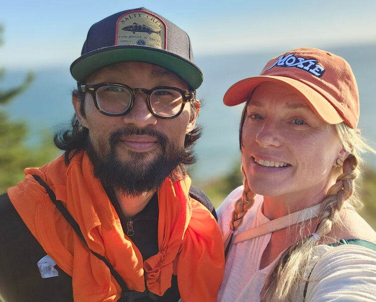 A middle age Korean man with round glasses and a beard, wearing a black hat with the image of a fish is posing next to a middle age white woman in braids and wearing an orange hat that says, "Moxie".