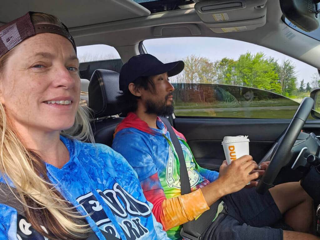 White woman and Korean man with a beard driving in a car. The man is holding a dunkin' coffee cup.