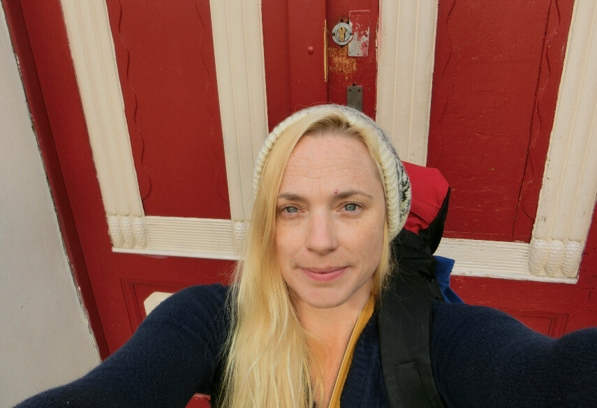 blonde woman with a backpack standing in from of a red and white door.