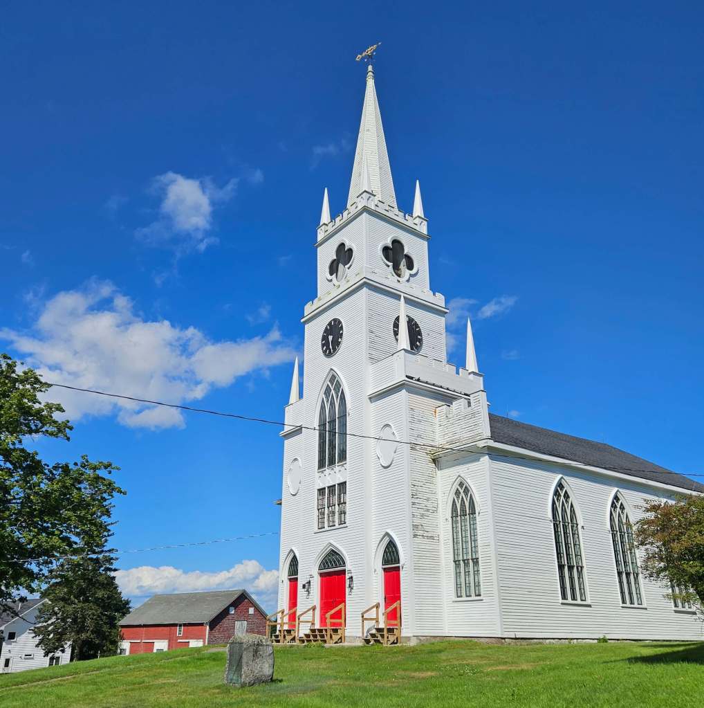 East Machias Congressional church. A tall white church with a single steeple built in the 1800's in Gothic Revival. Three bright red doors open to the church.