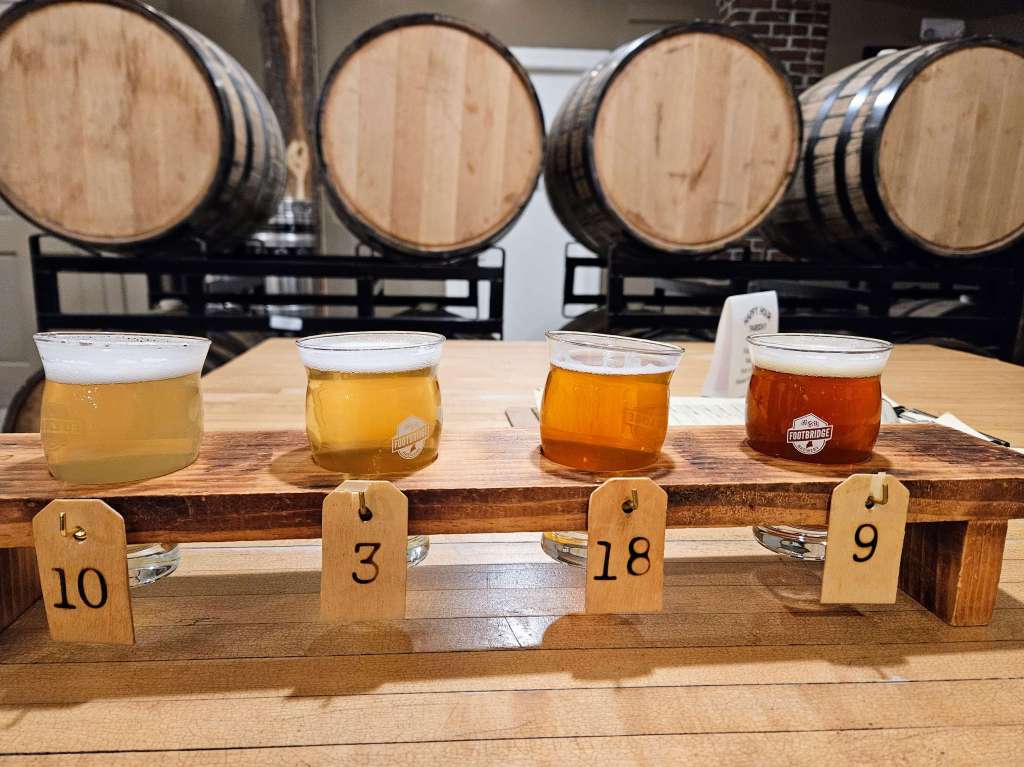 A flight of four beers each given a corresponding number. The numbers are from left to right: 10, 3, 18, 9. Behind the beers are wooden beer barrels.