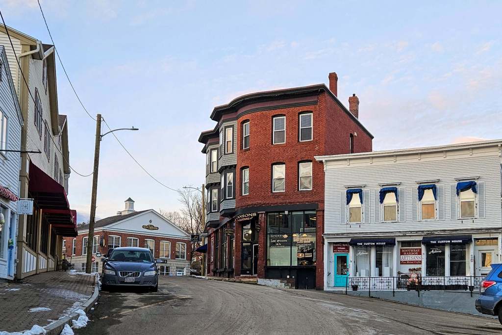 A curving downtown street with small store front building from the late 19th century. A tall three story red brick building stands out and above the other store fronts.