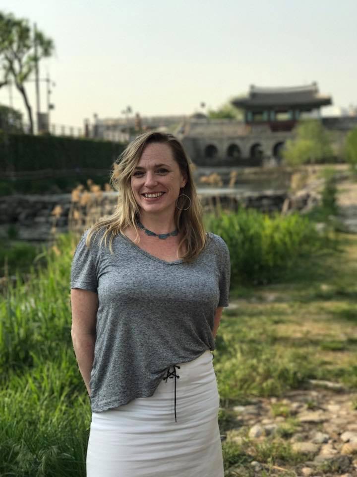Adrienna Ogin standing in front of Buksumun the northern floodgate of Hwaseong Fortress in South Korea. 
The season is spring and the scene is friendly and welcoming.